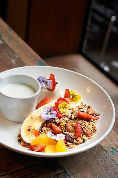 assiette petit déjeuner avec céréales banane, tasse de lait, fruits, panacotta, fraises et fleurs sur table en bois
